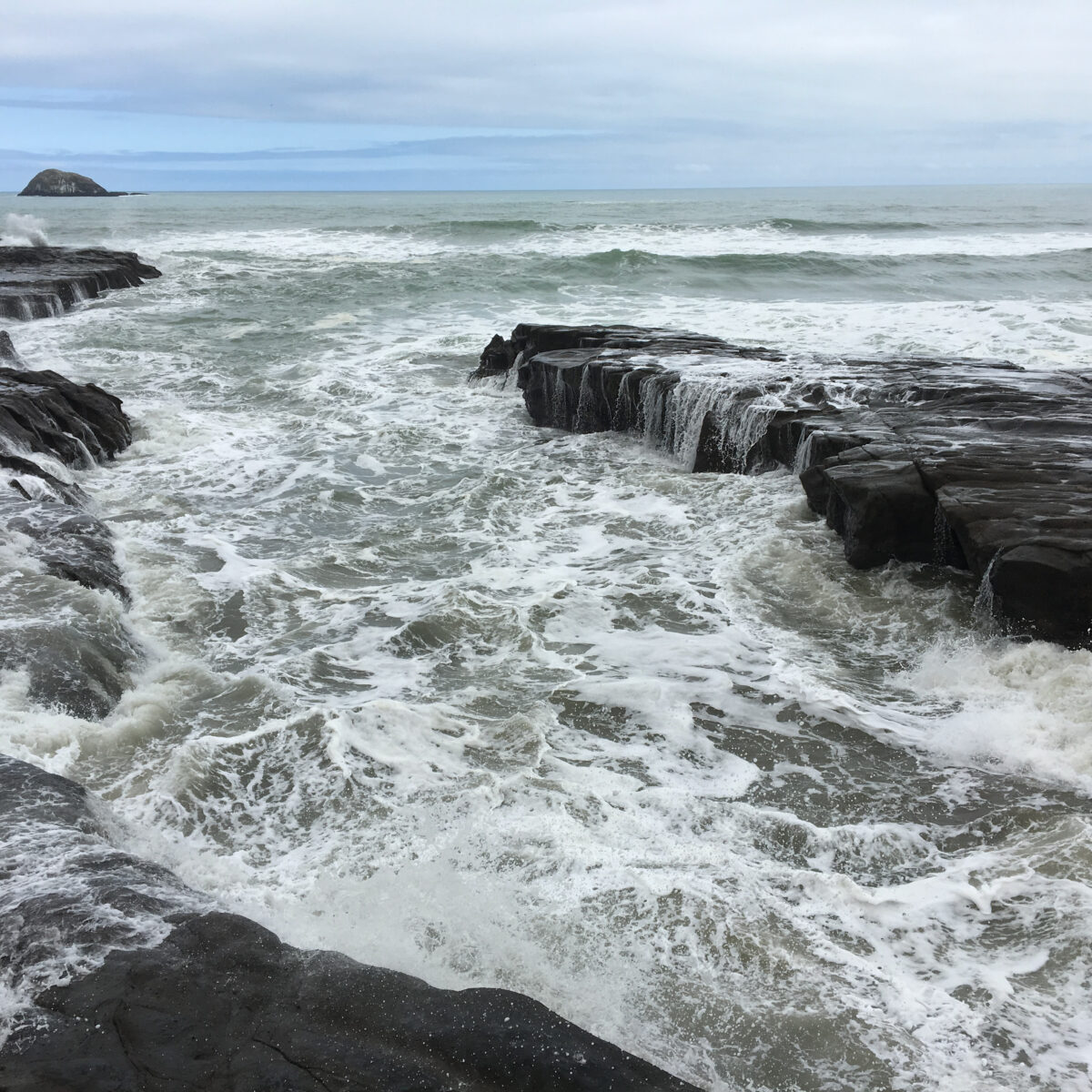 Rough sea crashes against dark layered rocks with white foam, a distant rocky island visible on the horizon under a cloudy sky.