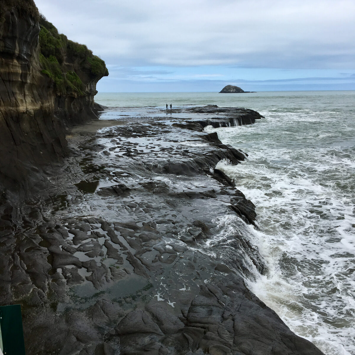 Cliffs with layered rock faces on left, tidal pools and rocky shore in foreground, two people standing near water, small island in distance.