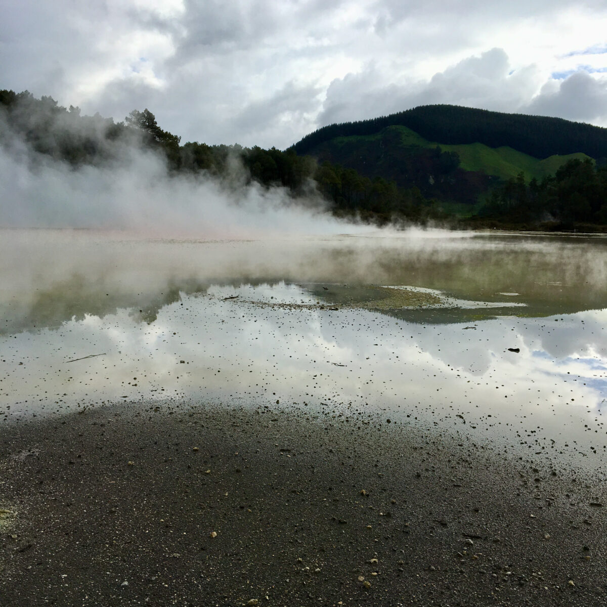 Mist surrounded lake with misty steam rising from water; distant green hills and dark forest under cloudy sky.
