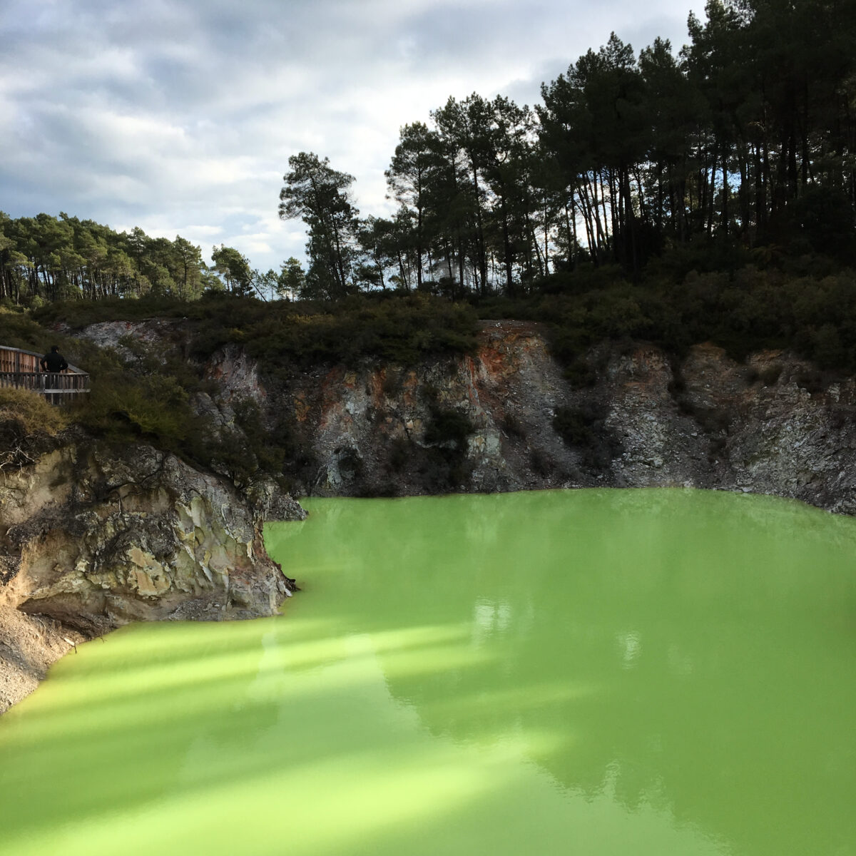 Green lake with rocky cliffside and dense pine forest in the background; a wooden viewing deck with a person stands on the left.