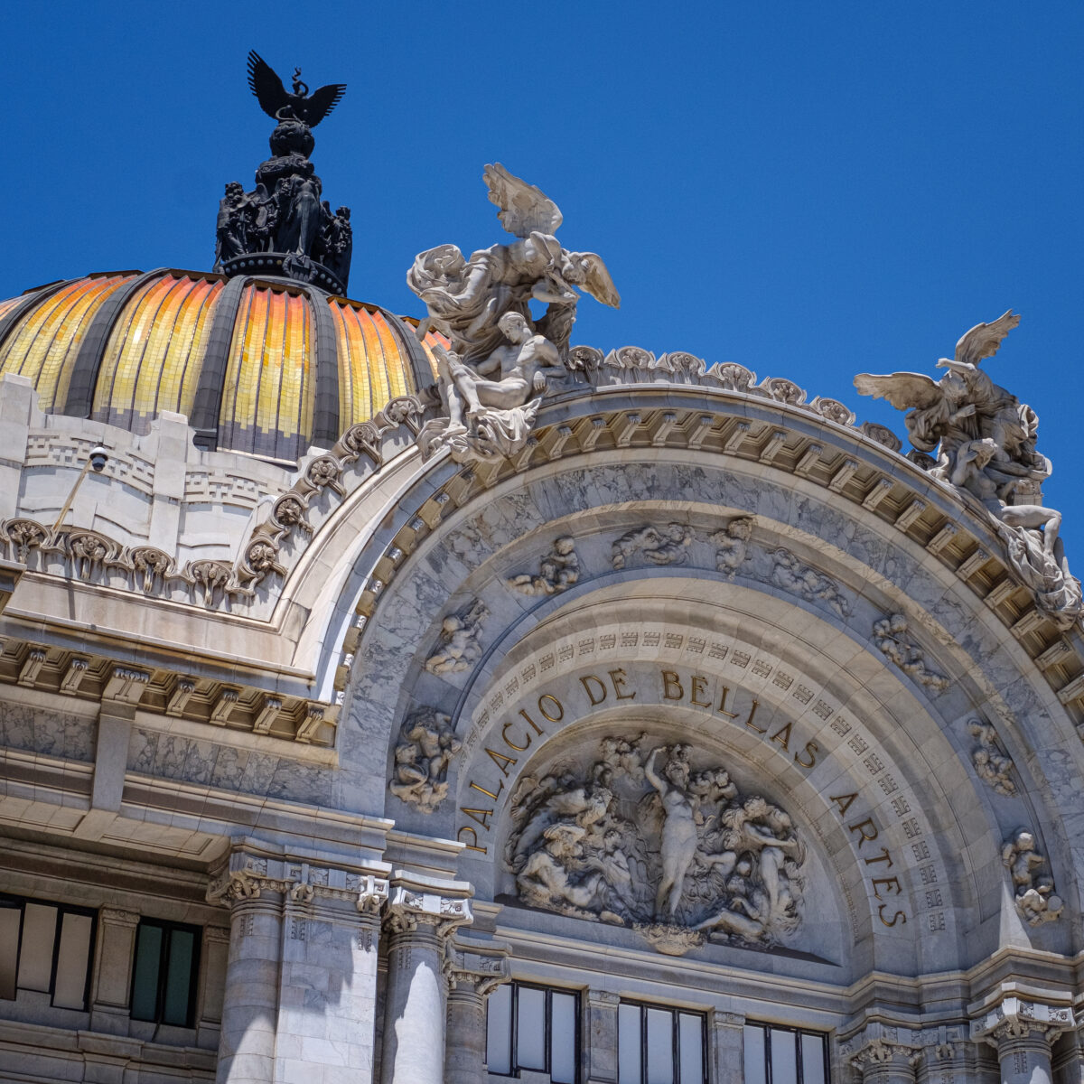 Facade of a grand neoclassical building with a colorful domed roof, ornate arches, and sculpted statues against a blue sky.