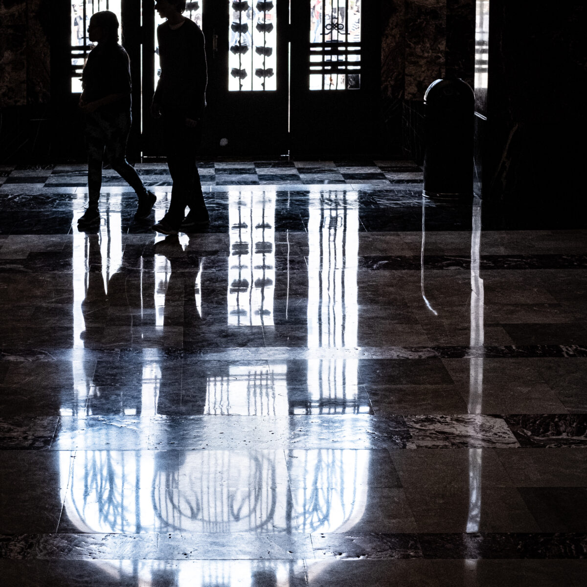 Silhouetted pair walks on a glossy checkered floor, reflected light from ornate glass doors creates vertical patterns.