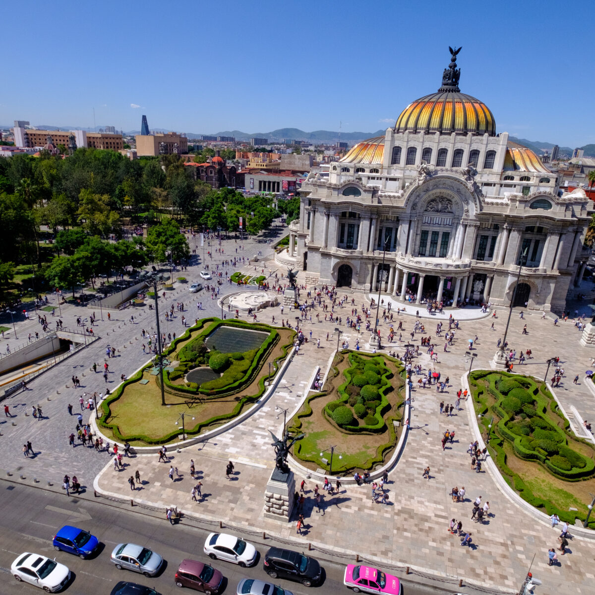 A grand neoclassical building with a colorful domed roof sits at the end of a wide plaza, surrounded by manicured gardens and crowds of people.