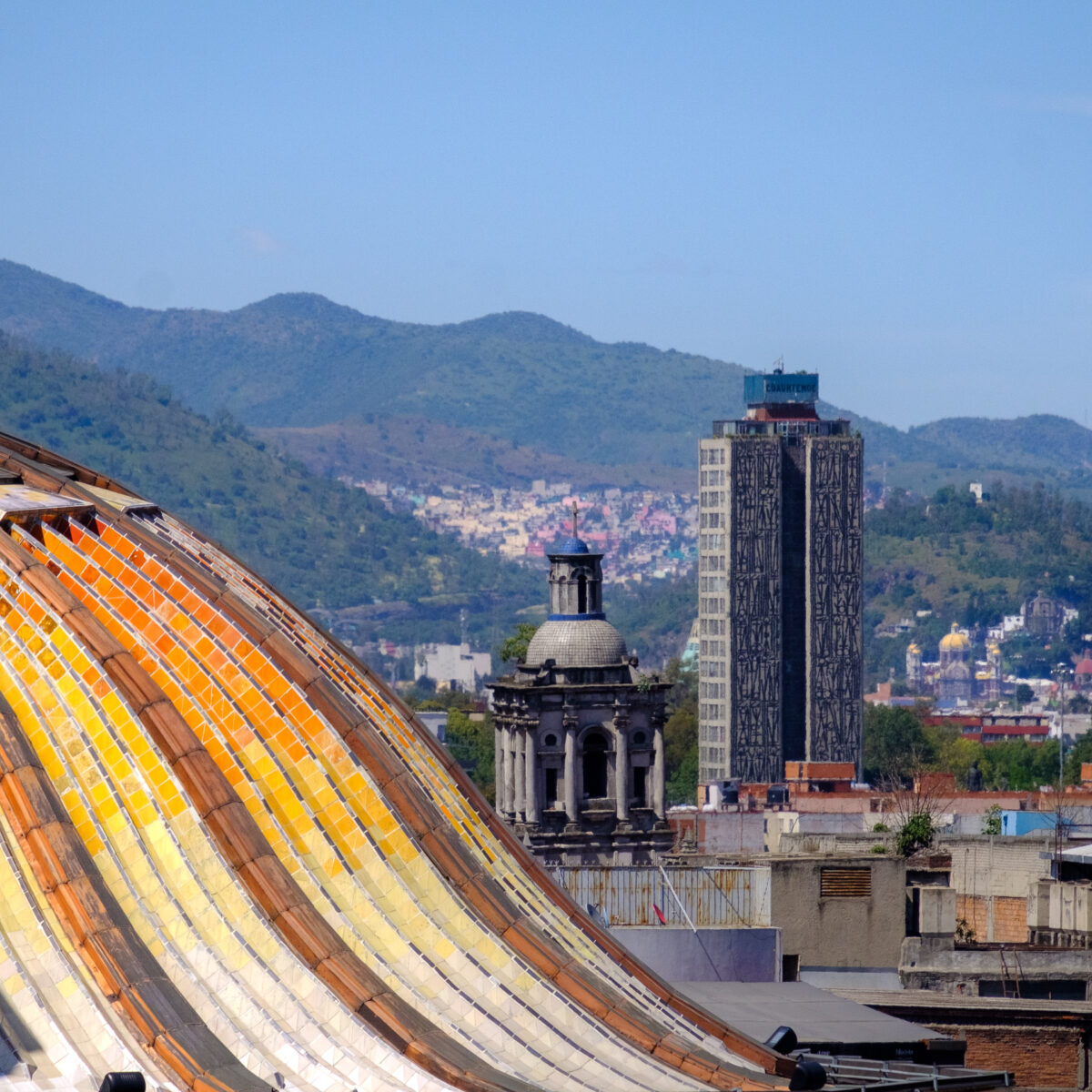 Glass-tiled domed roof with orange, yellow, and reflective strips in foreground; urban buildings and hills in distance under a clear blue sky.