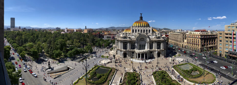 Panoramic view of a grand historic building with a yellow-orange dome, surrounding plaza, and bustling street traffic lined with trees on a sunny day.