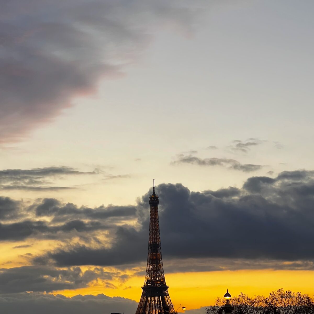 Eiffel Tower silhouette against a sunset sky with dark clouds and a glowing orange horizon, city lights at the base.