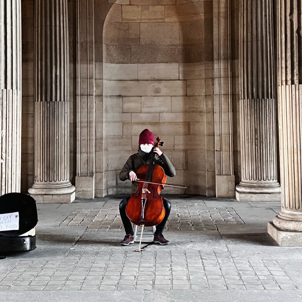 Cellist wearing a red hat and white mask, seated and playing a cello inside a stone colonnade with tall fluted columns.