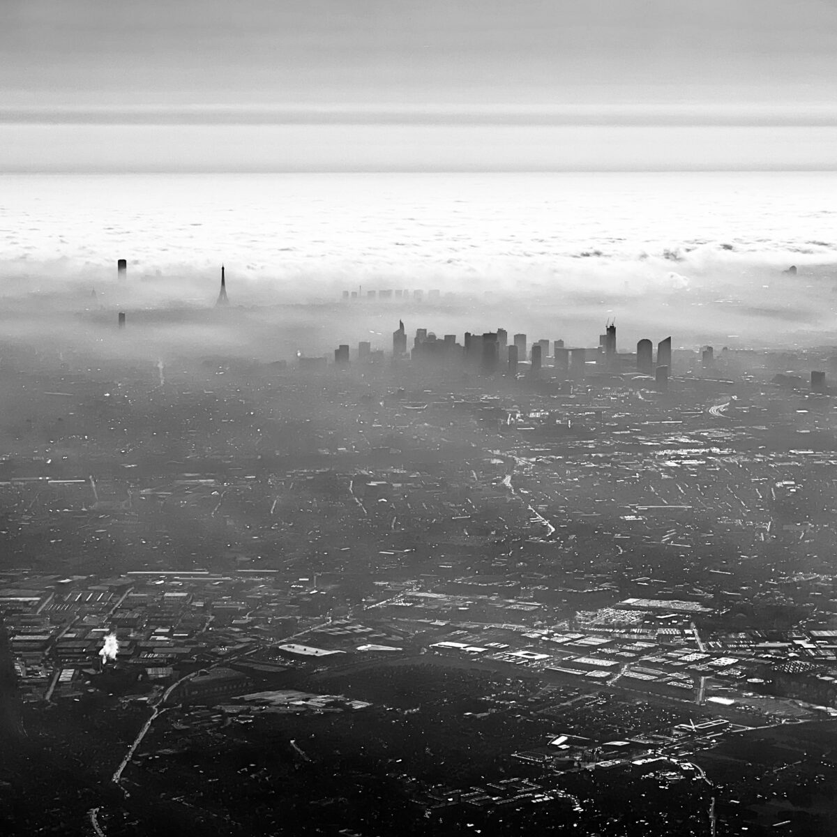 Aerial view of a city skyline emerging above low clouds, with tall buildings silhouette against a hazy horizon and a sprawling urban landscape below.