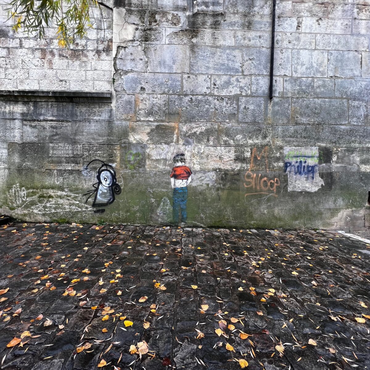 Graffiti of a person in a red shirt and blue jeans painted on a weathered brick wall, with scattered fallen leaves on a cobblestone ground.
