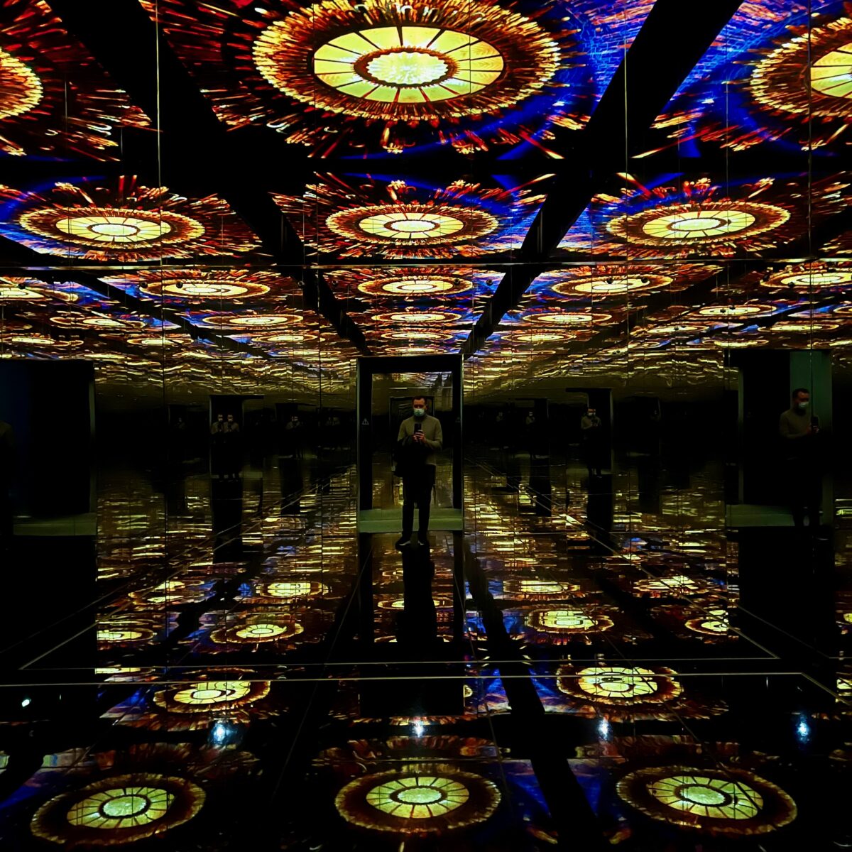 A person in front of a wall-to-wall mirrored room with circular stained-glass-like ceiling reflections and a reflective floor creating repeating patterns.