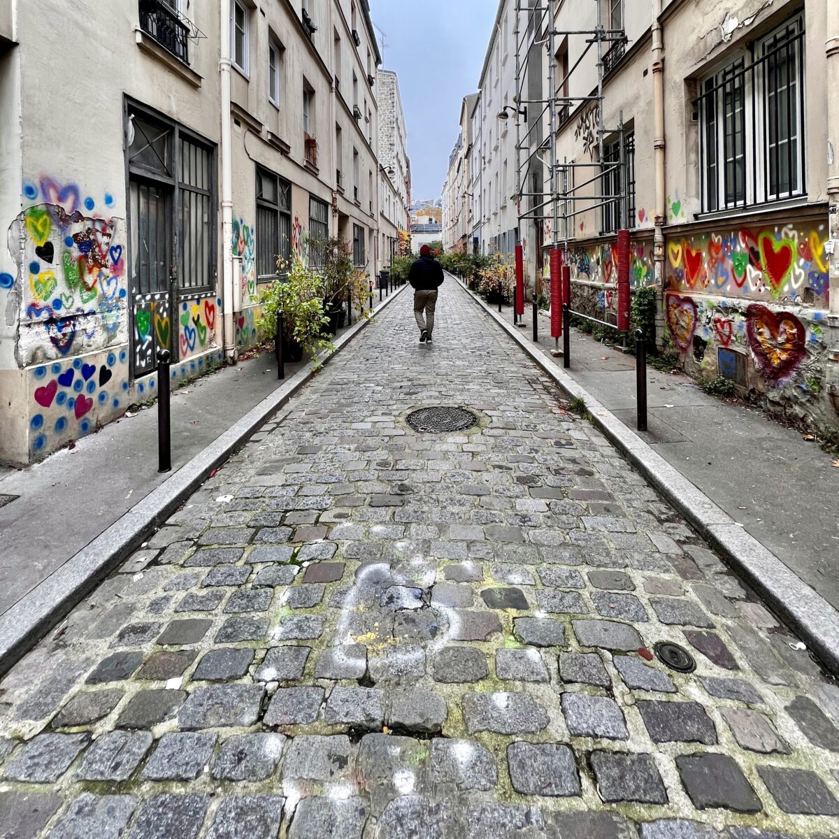 Cobblestone alley lined with graffiti-covered buildings, a single person walking away in the distance between two narrow sidewalks.