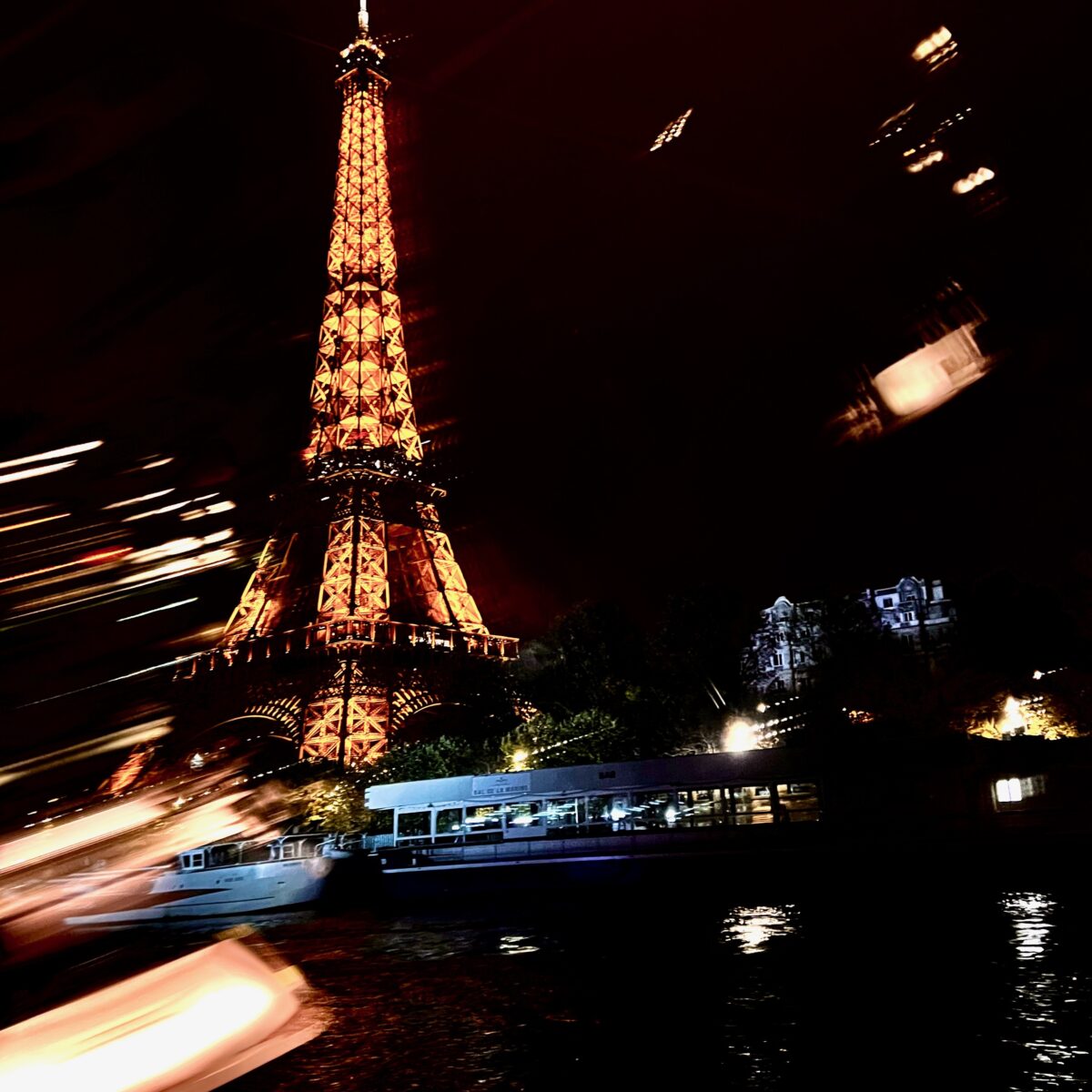 Eiffel Tower lit in warm orange at night beside the river, with blurred boat lights and a docked waterboat in foreground.