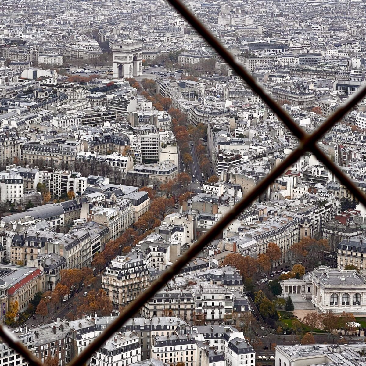 A dense Parisian cityscape viewed through a diagonal chain-link fence, with streets, Haussmann-style buildings, and the Arc de Triomphe in the distance.