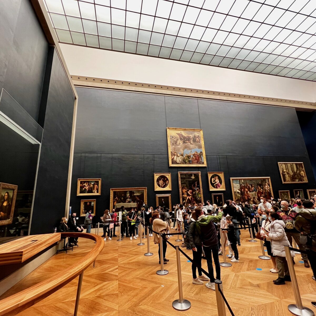 Crowded museum gallery with visitors lined up behind ropes, viewing large classical paintings on dark walls and a polished wooden floor.