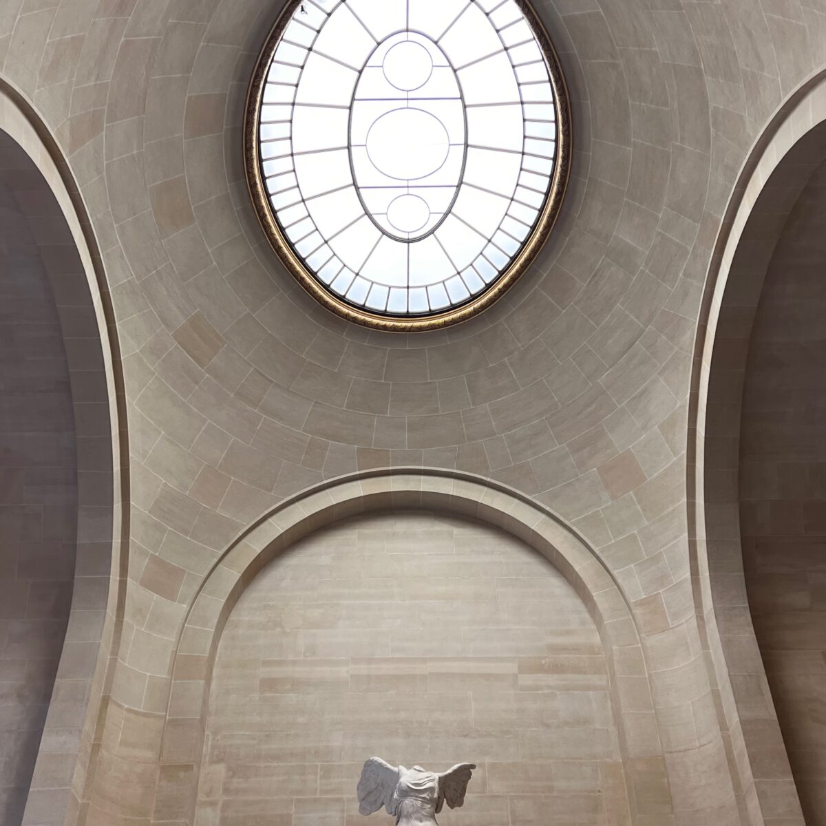 Interior view of a domed ceiling with a large circular skylight and a statue with wings at the lower center.