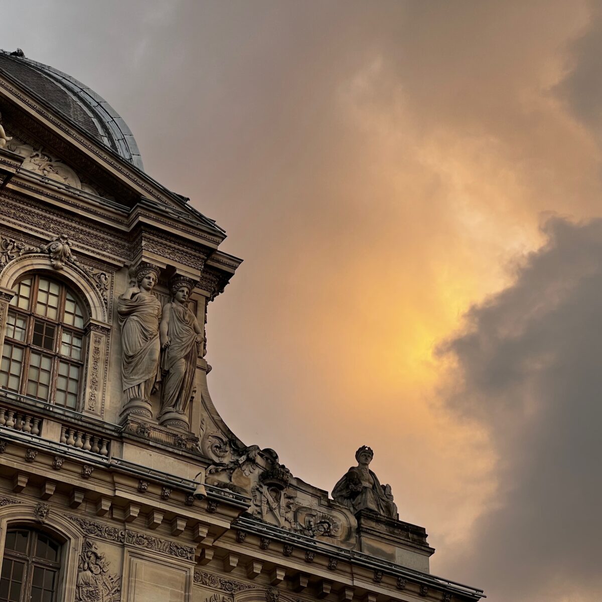 Ornate classical building facade with sculpted figures along a decorative cornice, arched window, and a dome against a sunset sky.