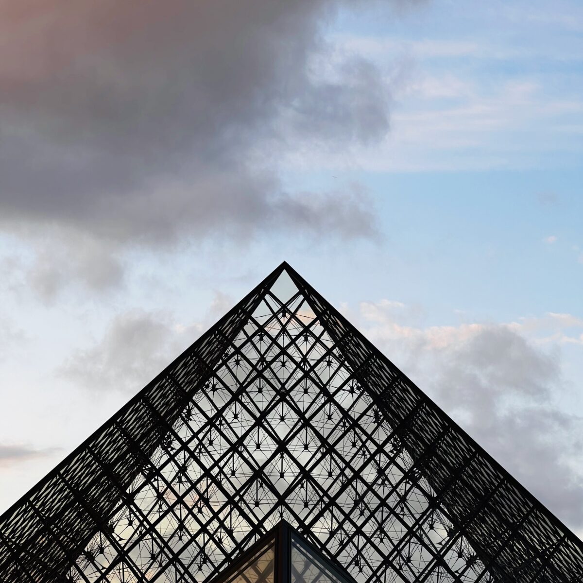 Glass and metal pyramid lattice against a partly cloudy sky, triangular peak at center with geometric grid patterns.