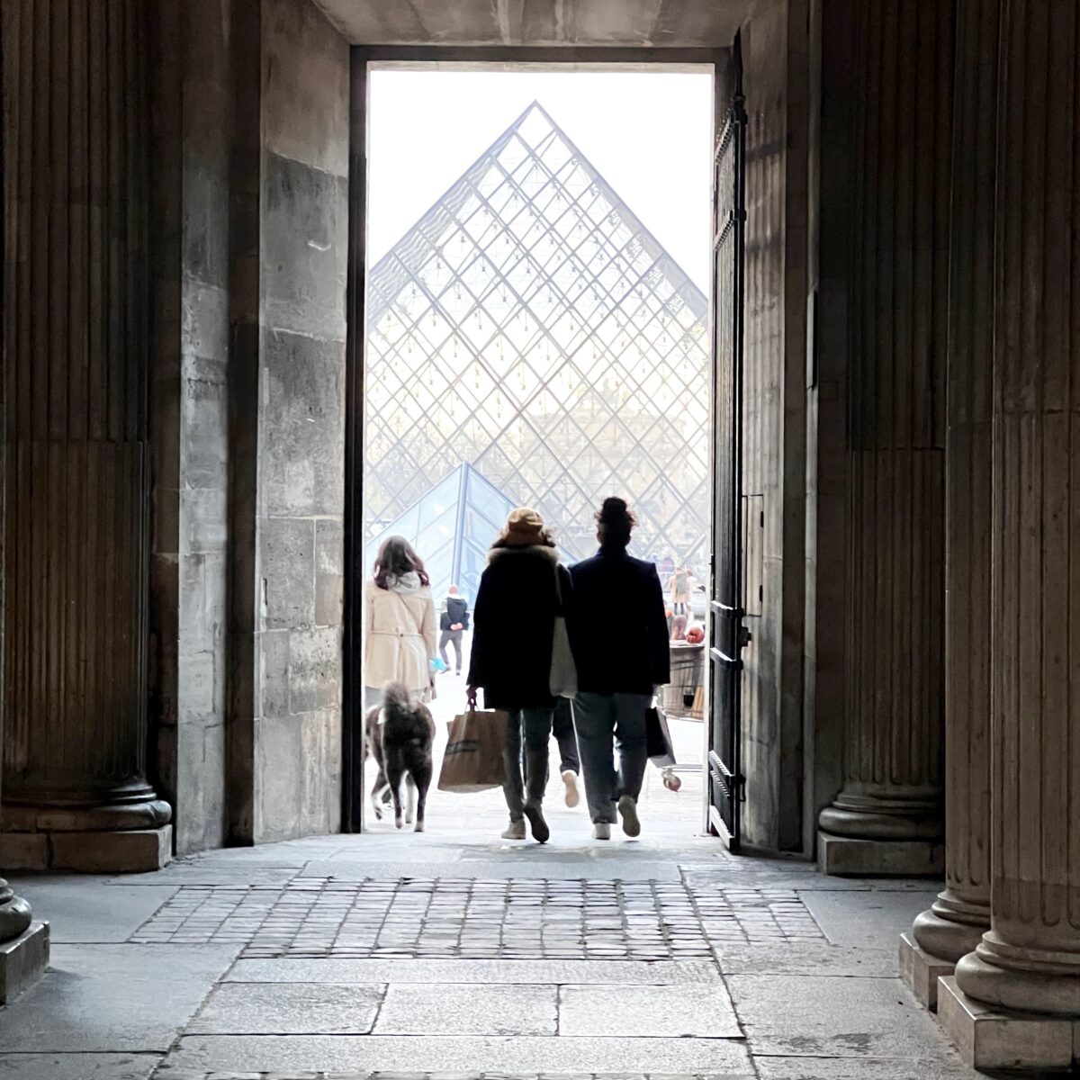 Two people walk out of a stone archway toward the glass pyramid at the Louvre, accompanied by a dog and shopping bags.