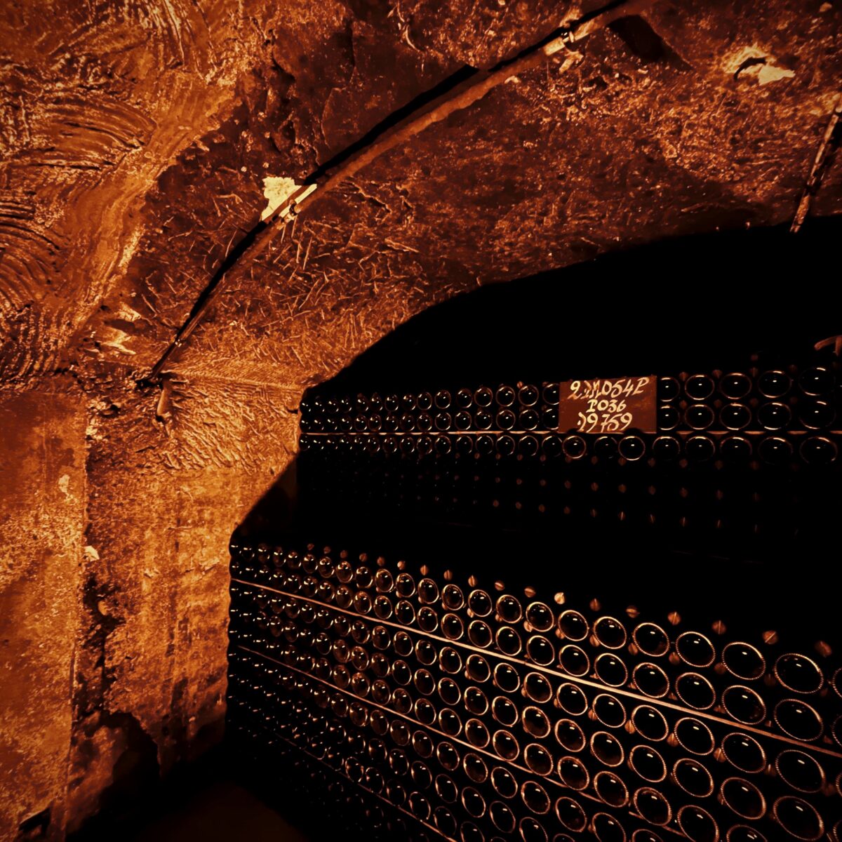 A dimly lit wine cellar with stacked bottles in a curved, rocky arched wall; a small sign showing number 36 is attached to the wall.