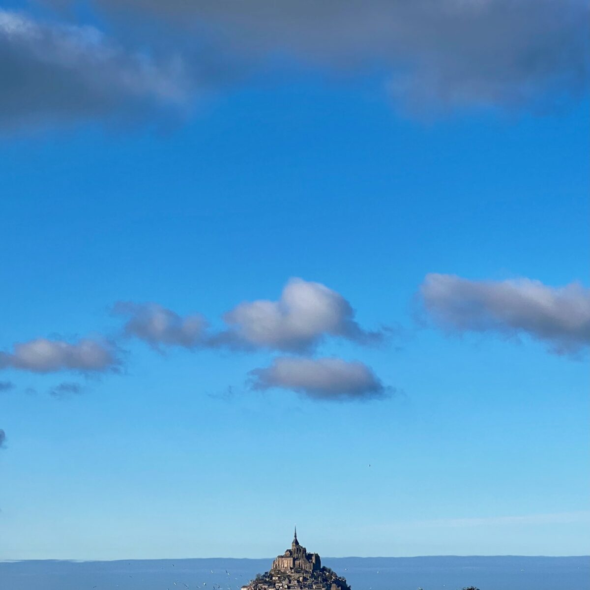 Mont Saint-Michel perched on a rocky island with surrounding sea, against a blue sky with scattered clouds, and a grassy foreground.