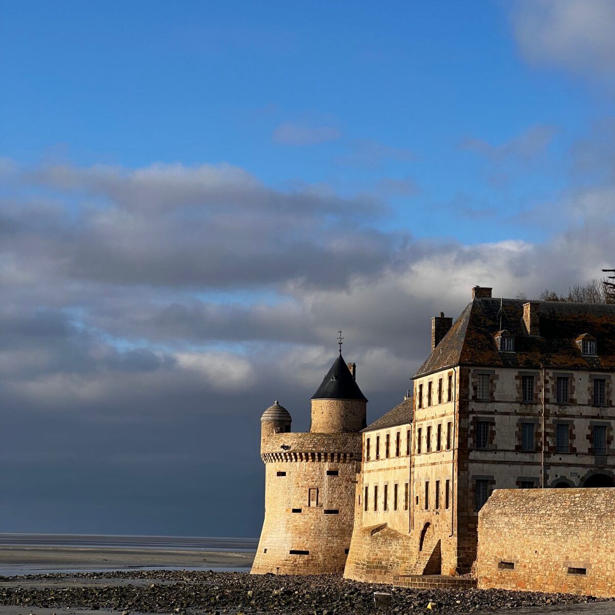 Fortified stone castle by the shore with round towers, attached rectangular buildings, and a rocky beach in the foreground under a blue sky.