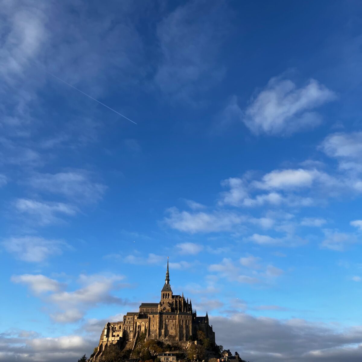 Mont Saint-Michel rises atop a rocky island with surrounding ancient stone walls and village buildings at its base, set against a blue sky with scattered clouds.