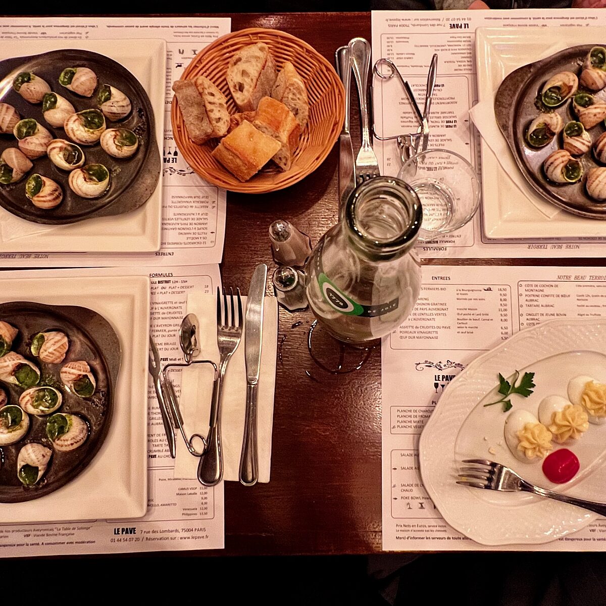 Several plates of stuffed deviled eggs with green garnish, a basket of bread, and a bottle of water on a dining table set with utensils.