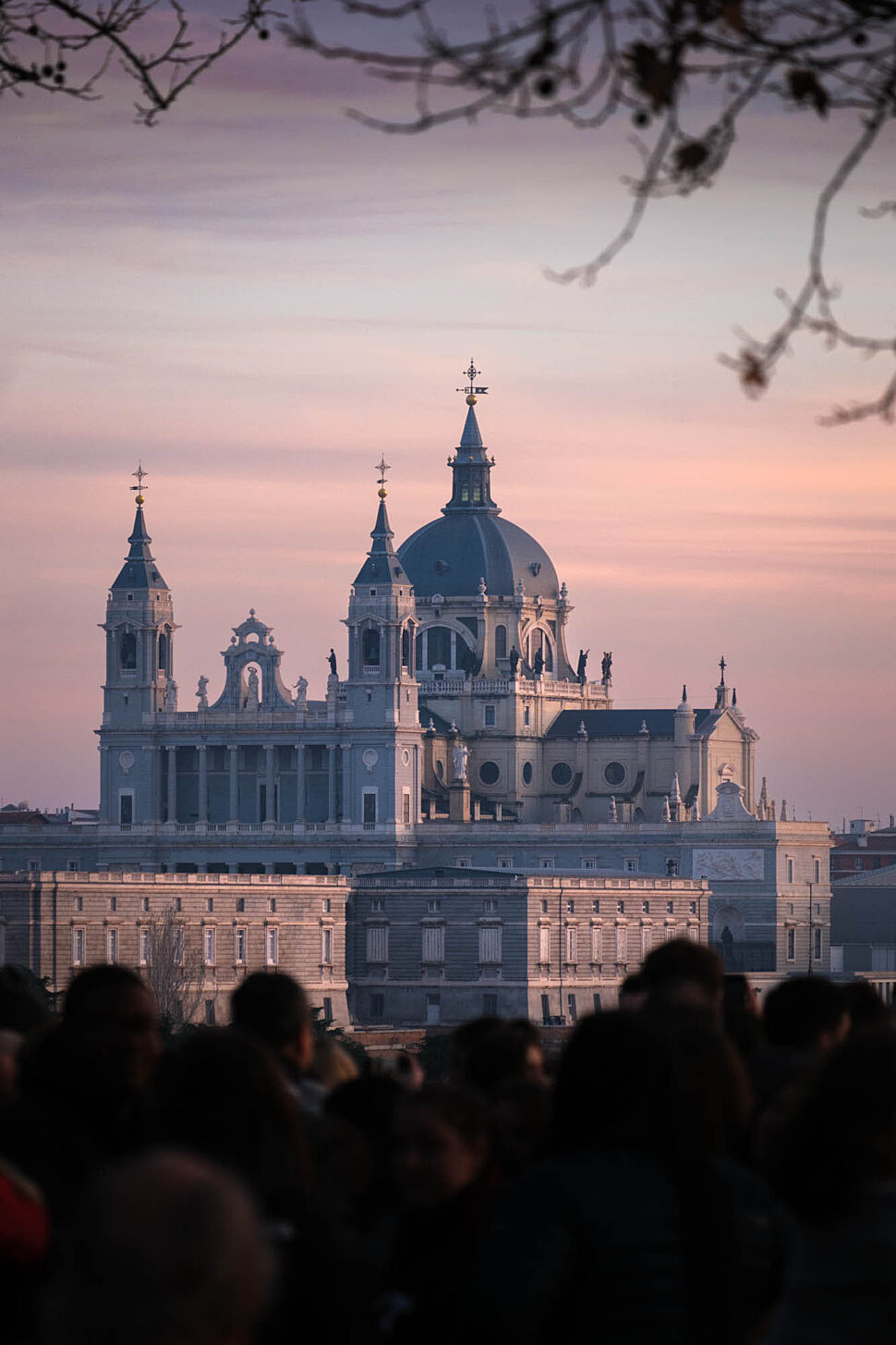 Dome-topped grand cathedral with ornate spires rises above a neoclassical facade, framed by silhouettes of people in the foreground at sunset.