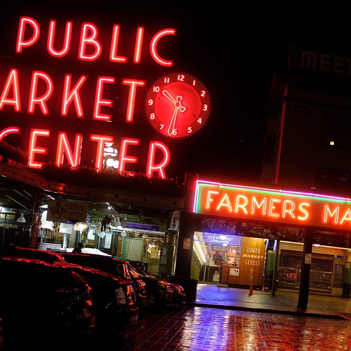 Night scene of Pike Place Market with large red neon "PUBLIC MARKET CENTER" sign and a bright "FARMERS MARKET" sign, wet pavement, and parked cars.