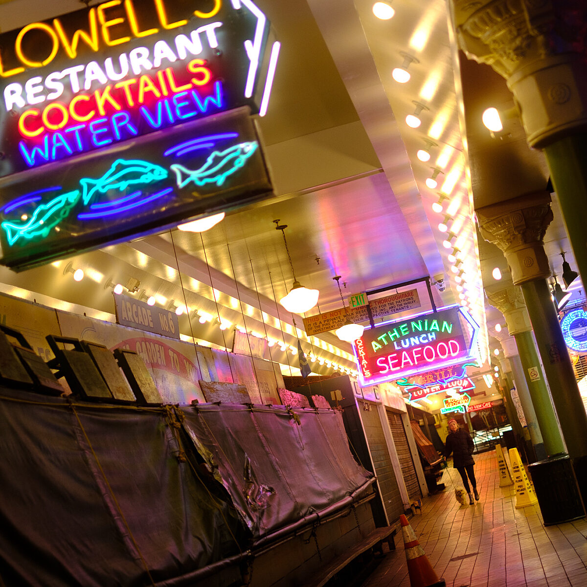 Nighttime city scene with neon signs for restaurants and seafood, along a lit storefront corridor with striped awnings and a person walking away.