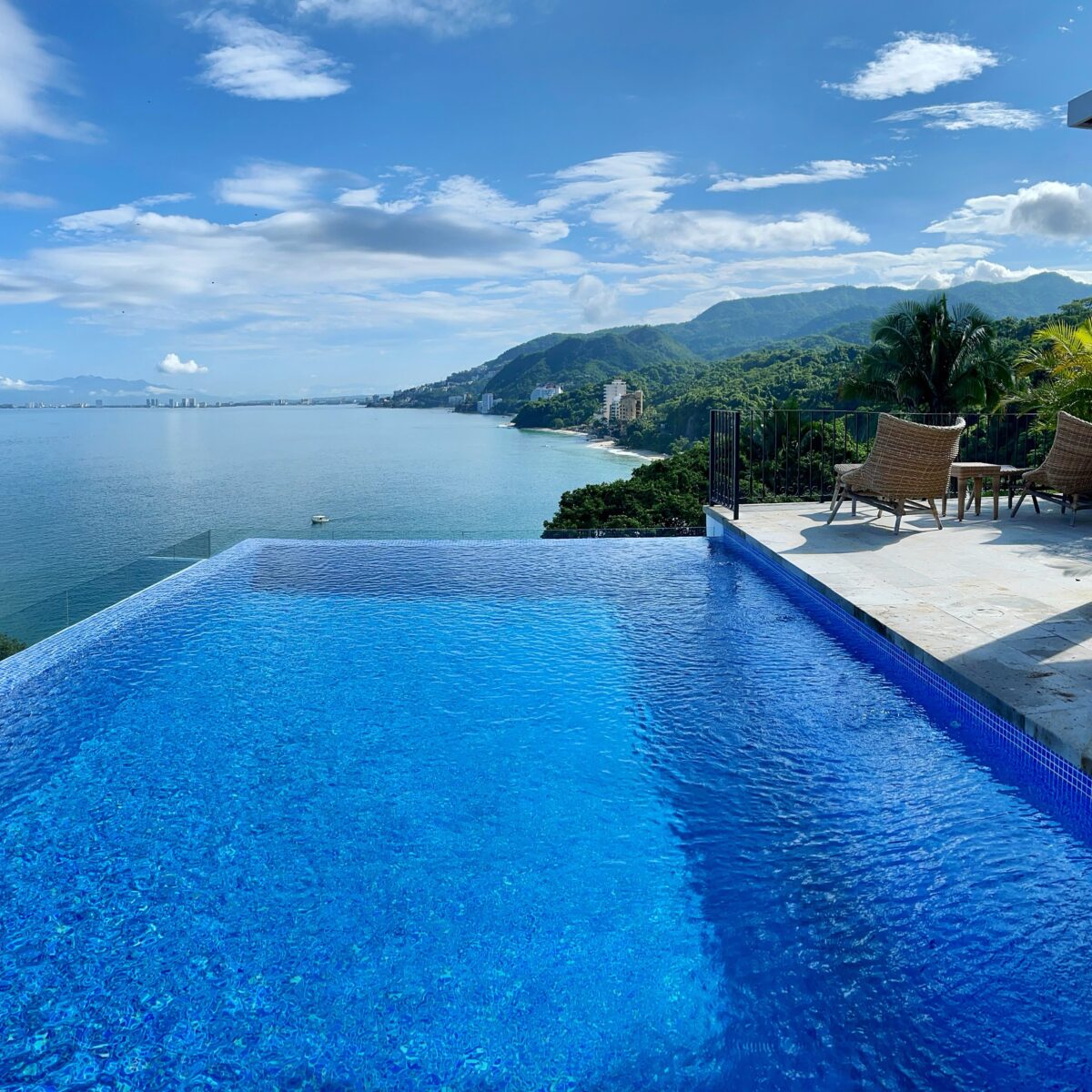 Infinity-edge swimming pool with blue tiles overlooks a calm ocean, hillside town, and mountains under a clear blue sky; poolside seating and leafy plants on the right.