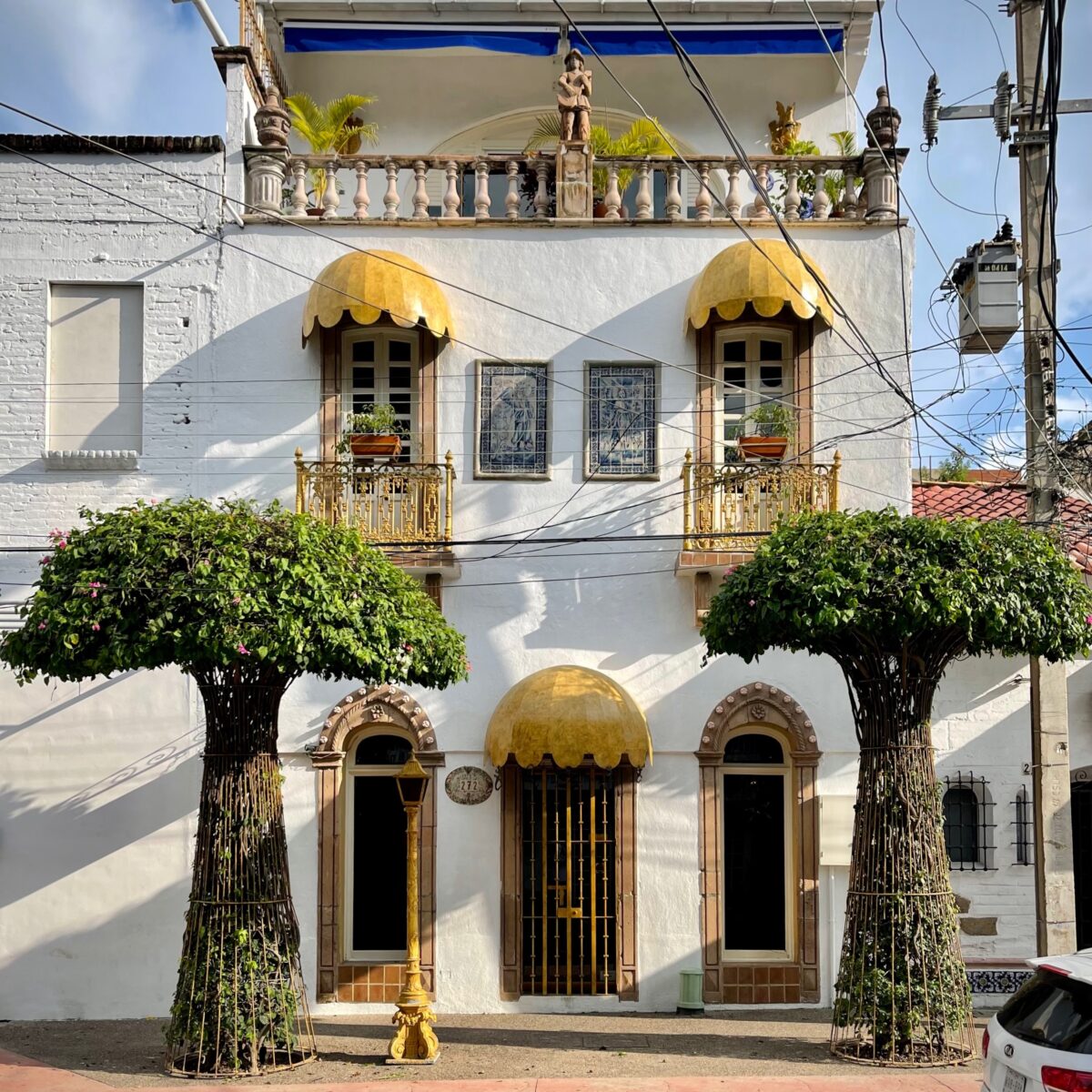 White two-story building with decorative gold awnings, arched doors, wrought-iron balconies, potted plants, and tree-shaped planters flanking the entrance.