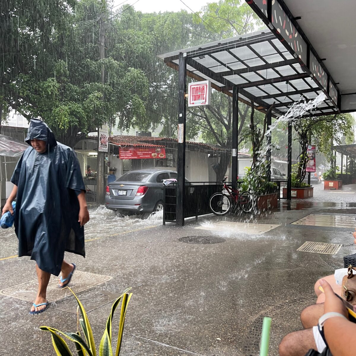Rain pummels a street view with a person in a blue poncho walking past a covered storefront as a car drives through puddles.