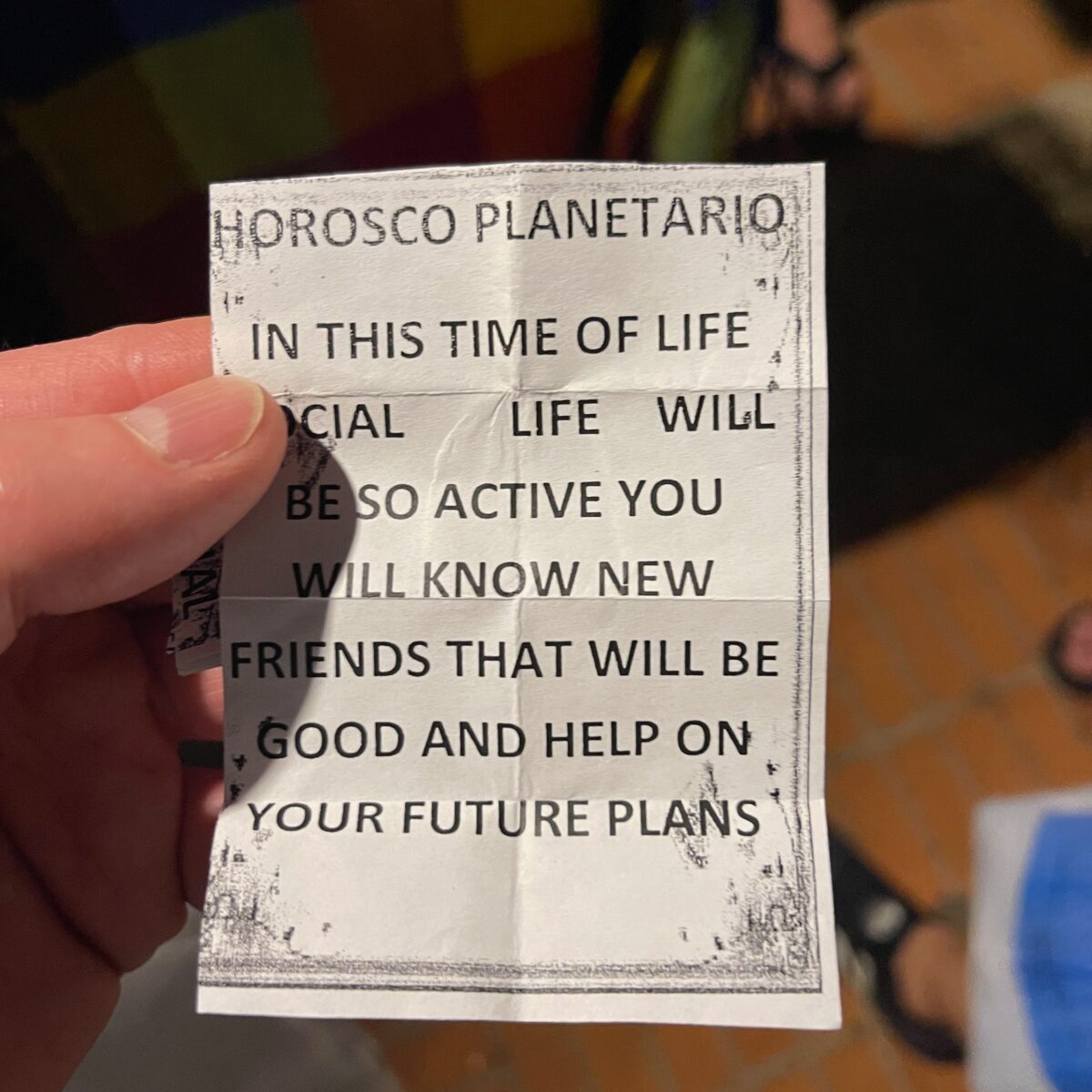 Small white fortune slip with black text reading "Horoscope Planetario" and inspirational message; held between fingers over a tiled floor.