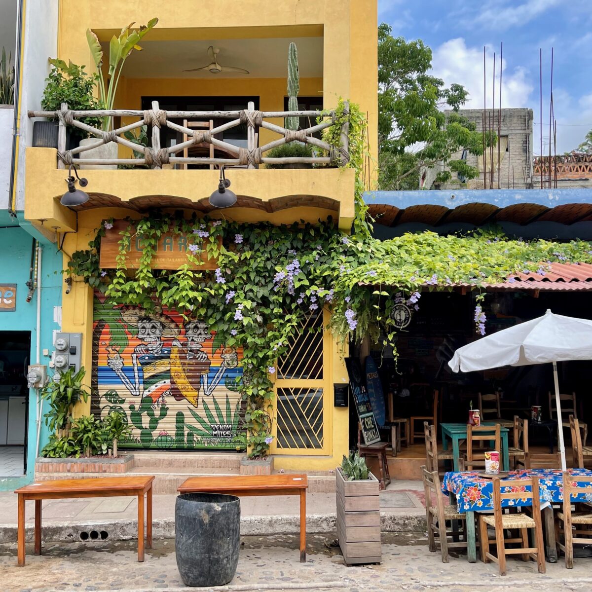 Colorful street scene with a yellow building, blue shop, and outdoor cafe seating under a white umbrella, vines and mural on a shuttered door.