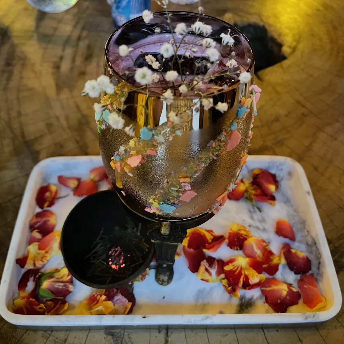 A metallic gold cocktail glass adorned with pastel confetti and white flowers sits in a tray of crushed ice and rose petals on a wooden table.
