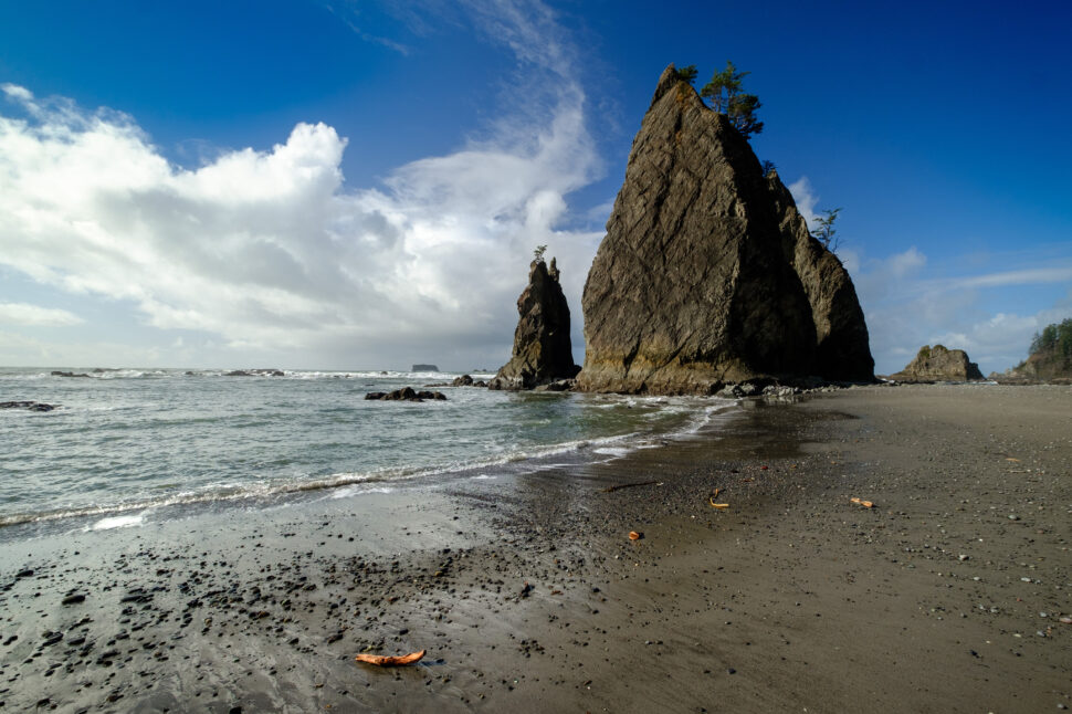 Two tall jagged rock formations rise from a rocky shoreline beside a calm sea, with scattered pebbles on a dark sandy beach and a blue sky with white clouds.