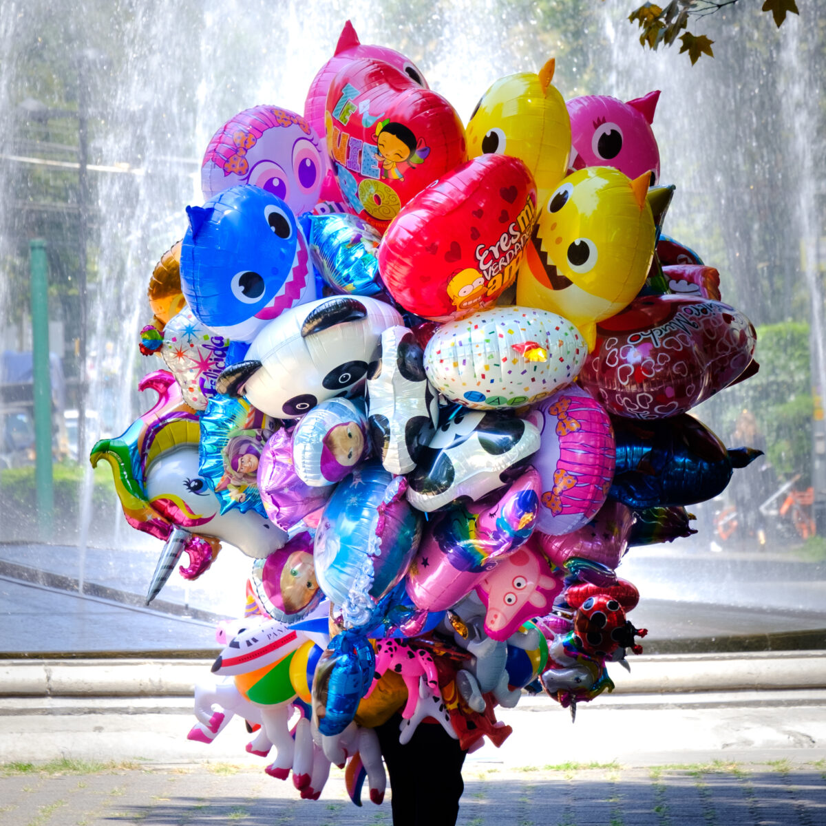 Person walking away with a large cluster of colorful helium balloons in various shapes and characters, in front of a water fountain.