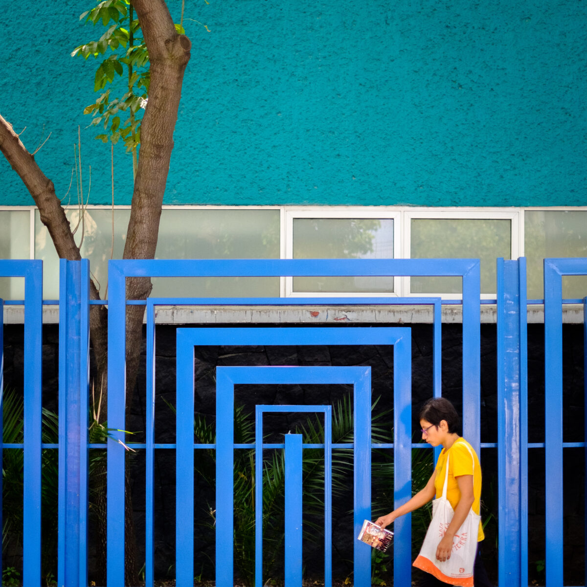 Person in yellow shirt walks past a bright blue geometric fence, carrying a tote bag and a booklet, with a teal wall and tree in the background.