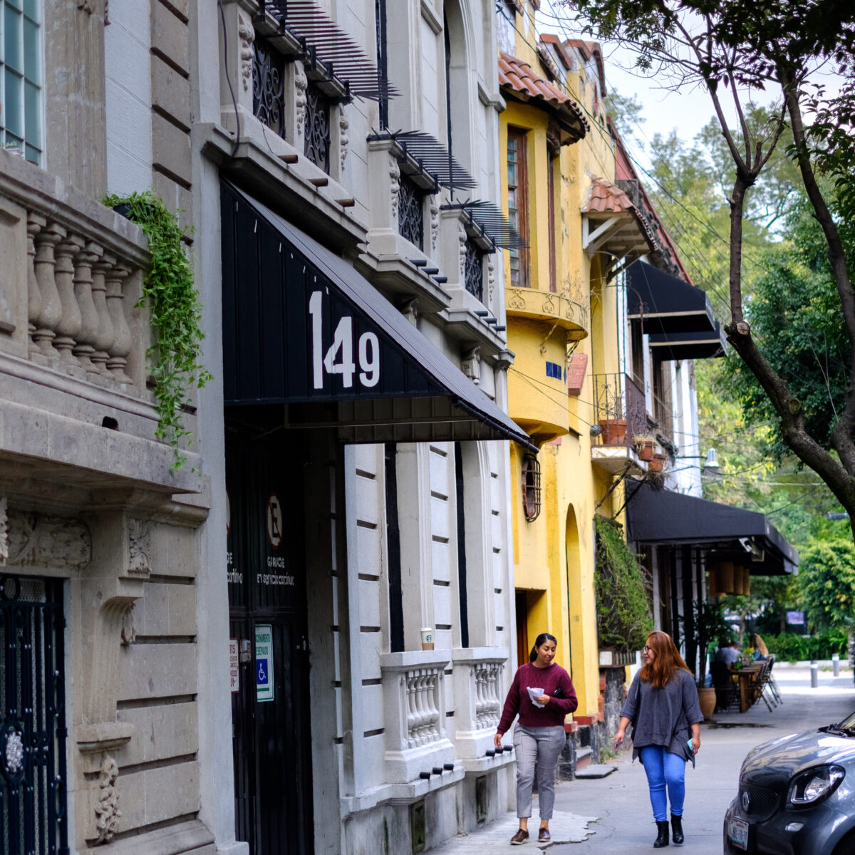 Two pedestrians walk along a tree-lined street past colorful, ornate buildings with a large "149" sign over a black awning.