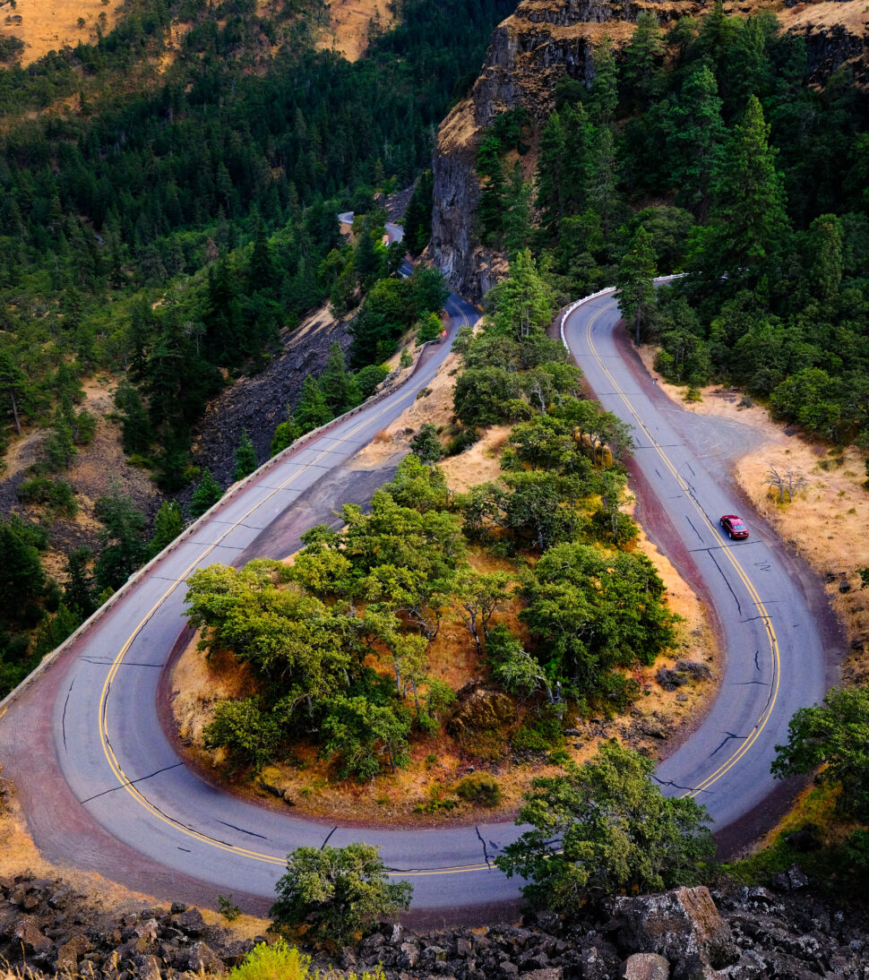 A winding mountain road loops around rocky cliffs, bordered by dense evergreen trees and dry grass, with a red car driving along the curve.