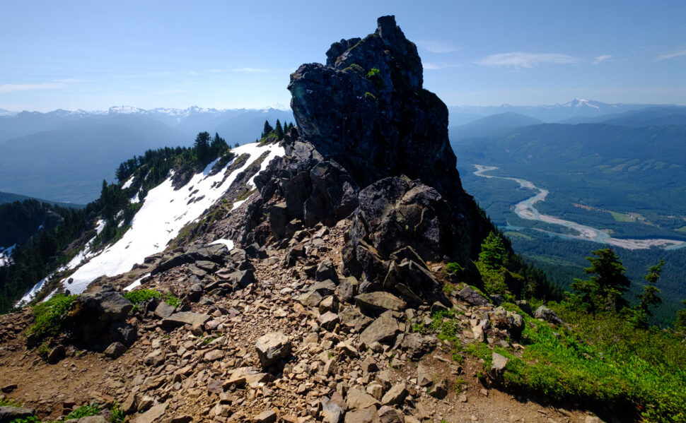 jagged rocky mountain summit with snow patches, green pines, and a winding valley below under a clear blue sky.