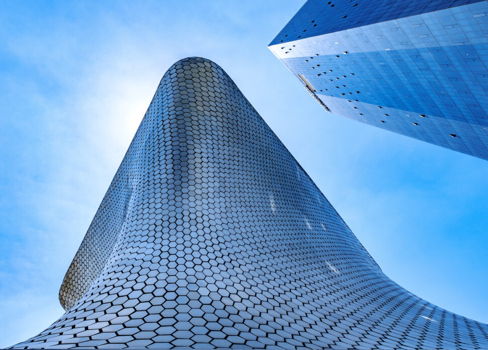 Curved metallic surface covered in hexagonal tiles meets a tall, angular glass skyscraper against a bright blue sky.