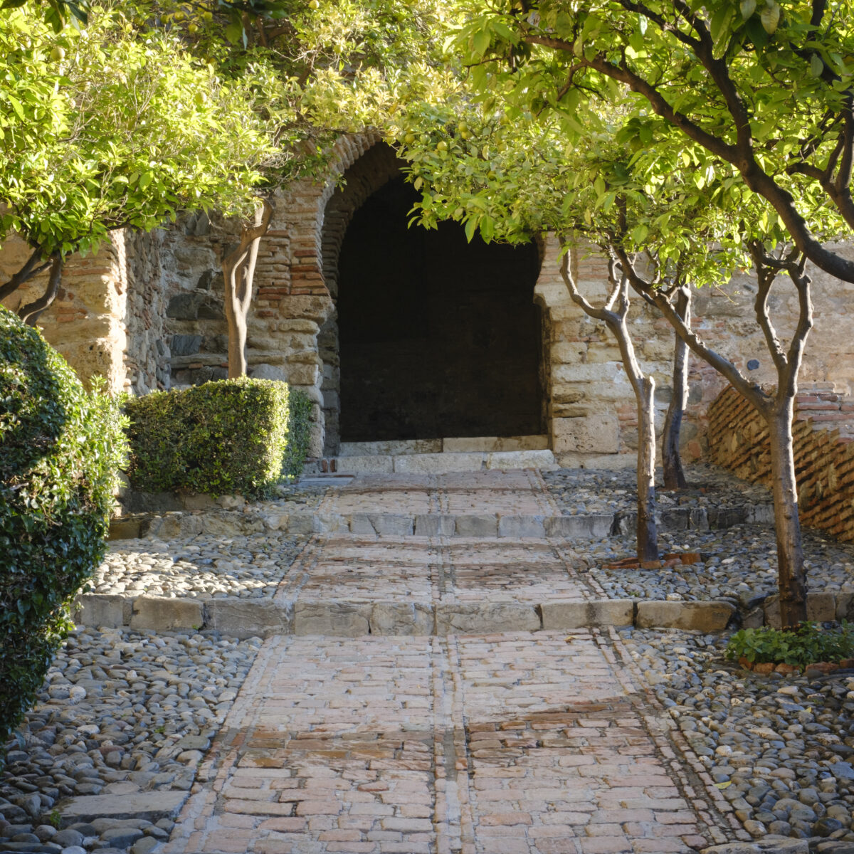 Stone pathway with brick steps leads to a dark arched doorway, framed by brick walls and lush green trees and shrubs.