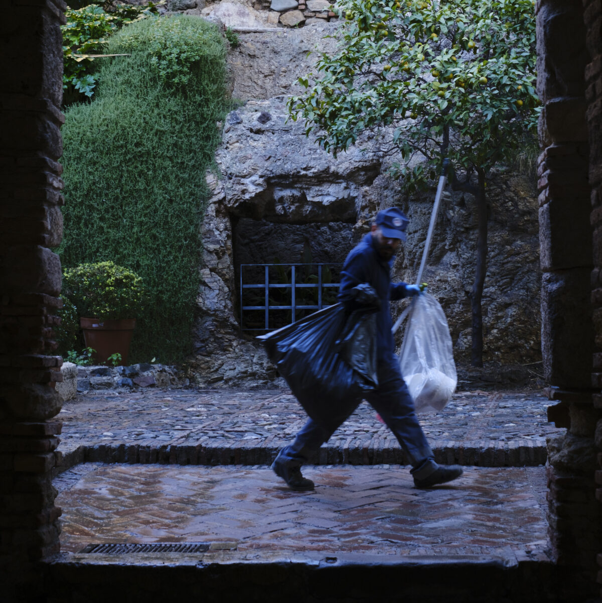 Man with blue uniform carrying two large trash bags walks through a stone courtyard framed by archways, with greenery and a lemon tree nearby.