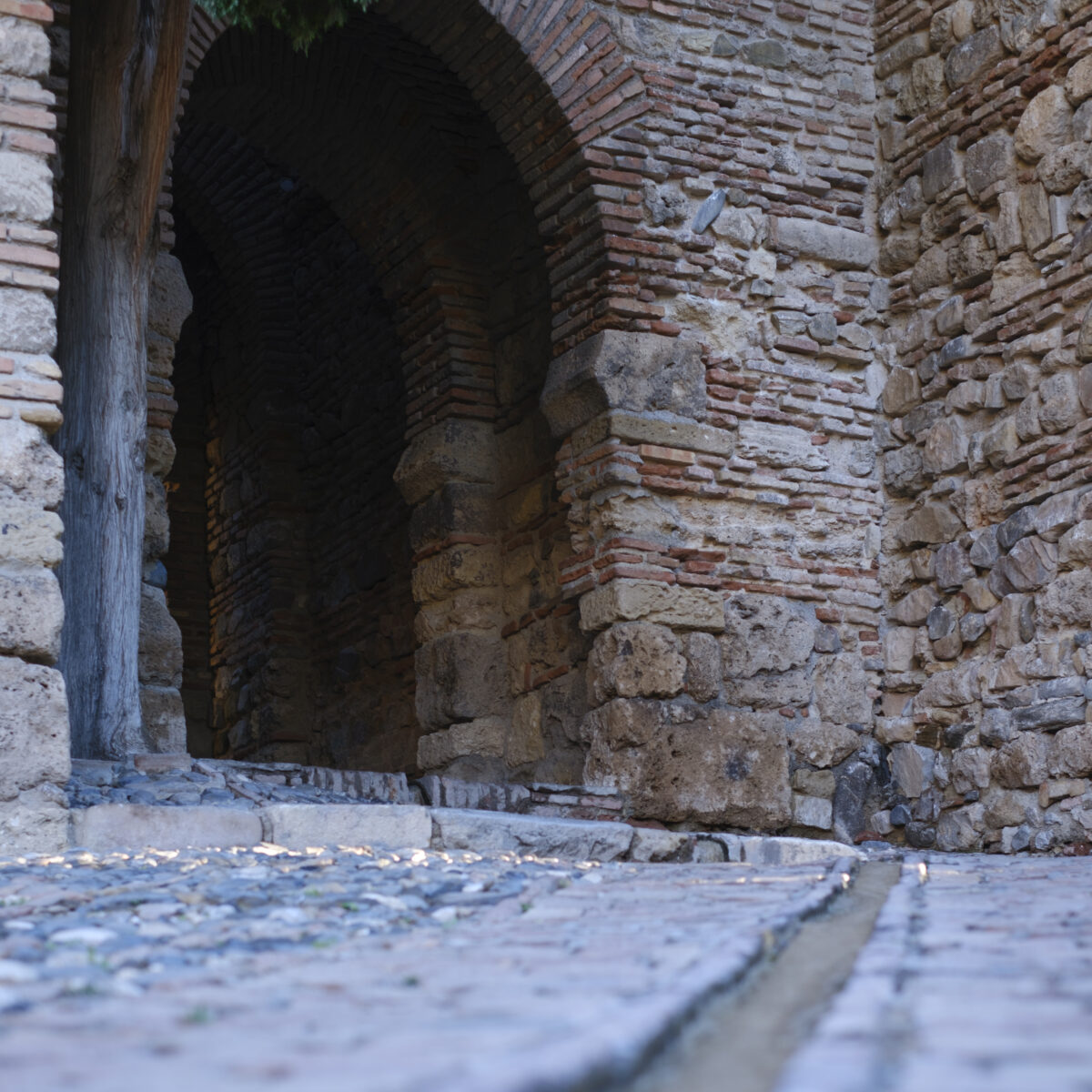 Old stone archway and brick walls lining a cobblestone path leading into a dark tunnel.