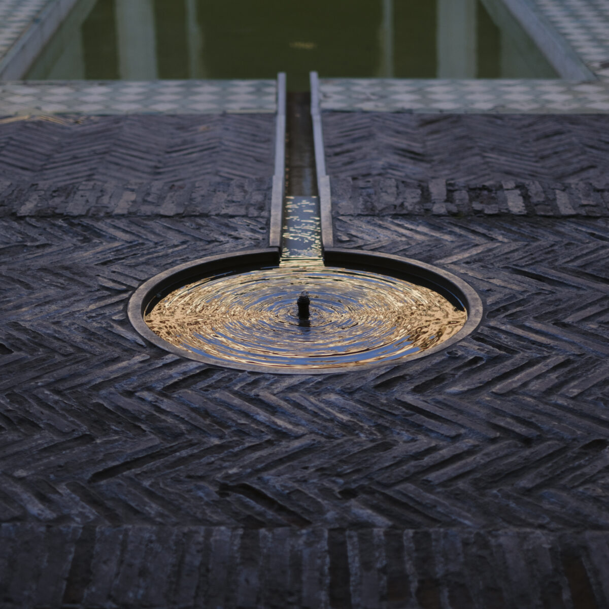 Small circular pool of rippled water at center, set in dark textured pavement, flanked by narrow metal channel leading to a green reflective pool.
