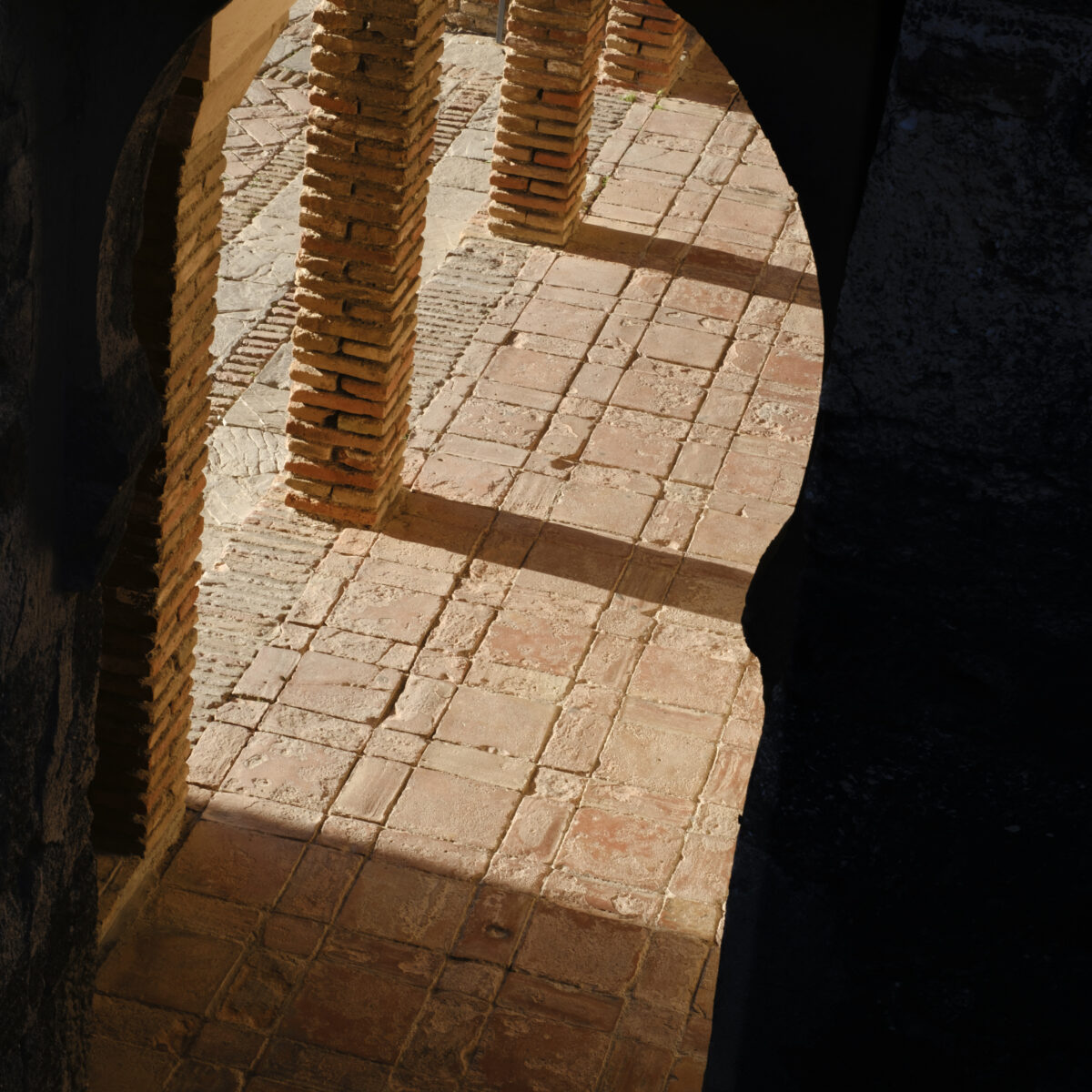 Sunlit brick arches and tiled floor seen through a dark arched opening, with stacked brick columns casting shadows on the pavement.