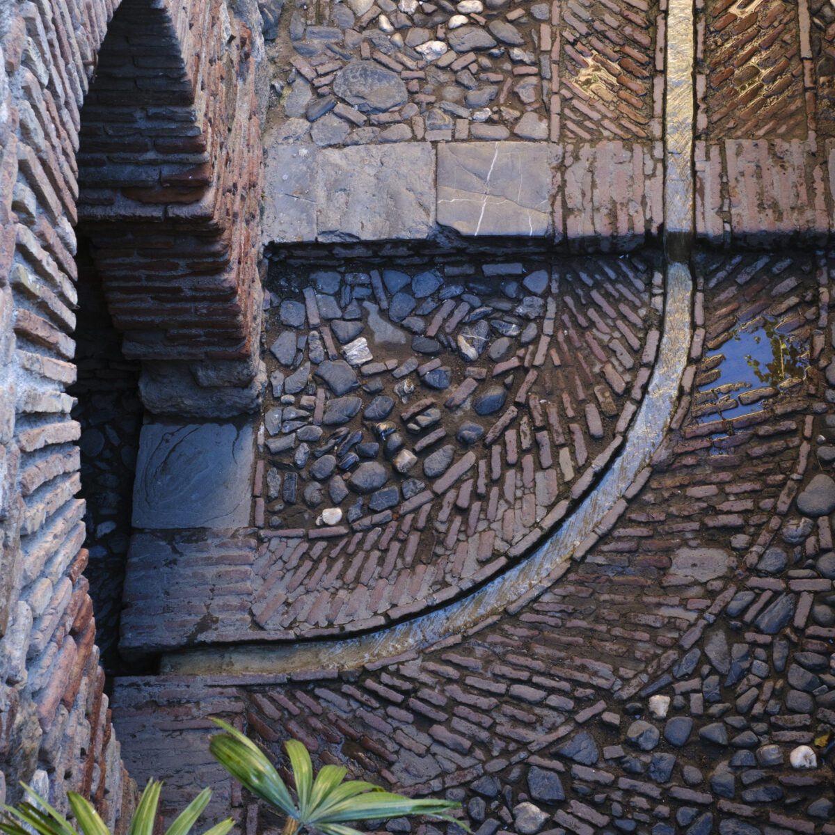 Curved brick and stone stairwell with a circular stone pattern, surrounded by red brick walls and small palm leaves in the foreground.