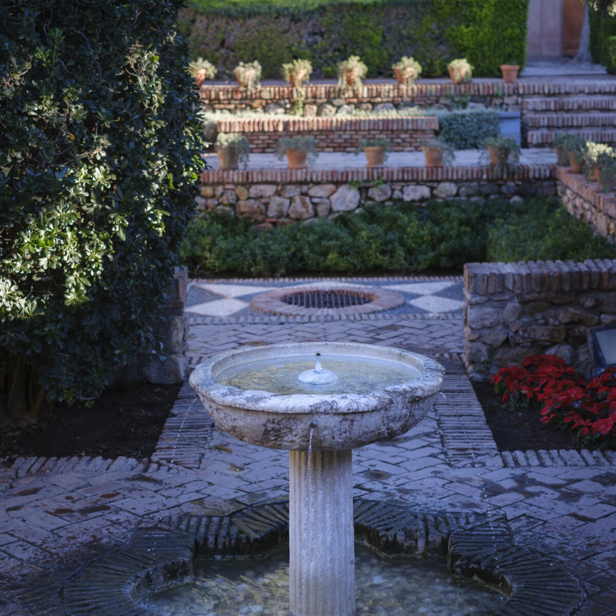 Stone courtyard with a weathered circular fountain in the foreground, brick terraces with potted plants, and trimmed hedges in the background.